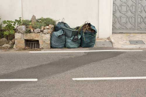 A row of four large, dark green refuse bags filled with various types of waste material, positioned on the edge of a pavement next to a cream-colored wall with a small garden bed containing rocks and plants. The bags are partly leaning against a decorative metal gate on the right side of the image. The scene appears to be in an urban residential area, with the bags placed on the street for removal. The pavement and street surface are visible in the foreground, with white painted road markings. The overall setting suggests a rubbish removal service, such as those offered by Office Clearance Bethnal Green, operating in the Bethnal Green area of London, with the scene illustrating typical street-level waste collection designated for rubbish clearance.