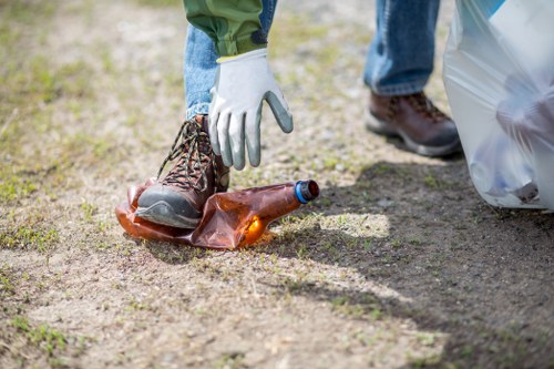 A person wearing white gloves and brown work boots is seen picking up a small pile of rubbish from a dirt surface outdoors. The rubbish includes a brown glass bottle partially filled with liquid, lying on its side, with an orange-brown cap attached. In the background, another pair of legs wearing jeans and dark shoes is visible, along with a large clear plastic bag that appears to contain additional waste. The setting suggests an outdoor area that may require rubbish removal services, with natural ground conditions and minimal vegetation present, indicative of a temporary or maintained space. This image illustrates the process of waste collection or cleaning, which aligns with rubbish removal activities carried out by companies such as Office Clearance Bethnal Green, supporting efforts to manage debris and unwanted items effectively in local areas near Bethnal Green, London.