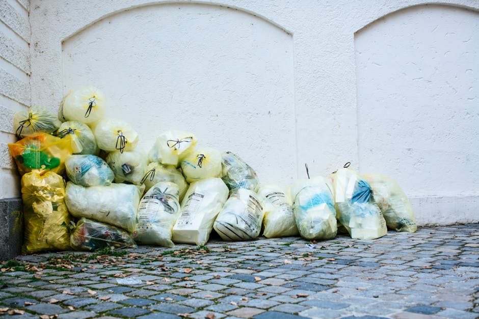 A row of five purpose-built waste bins lined up against a white exterior wall, situated on a paved surface with a low brick wall behind them. The first bin on the left is bright yellow with a lid slig