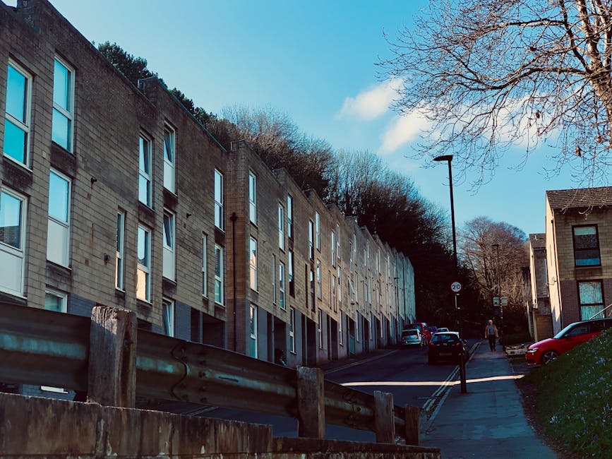 A row of older, semi-detached brick houses with white window frames and red-tiled bay windows situated along a quiet residential street. In the foreground, a parked silver car is aligned next to the c