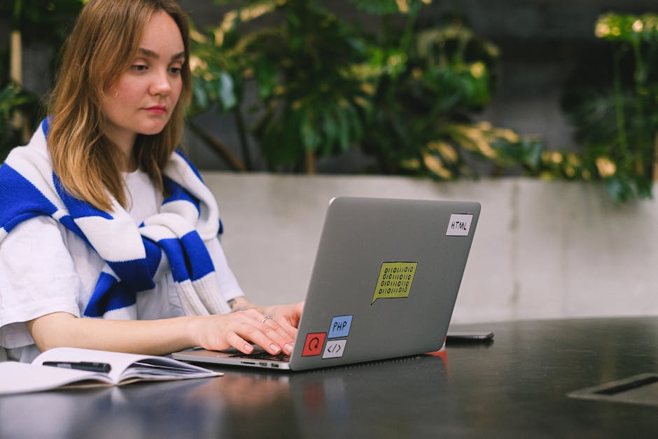 A person wearing a striped long-sleeve shirt is working on a laptop computer displaying lines of code, placed on a white desk. The individual’s right hand is on the keyboard while the left hand rest