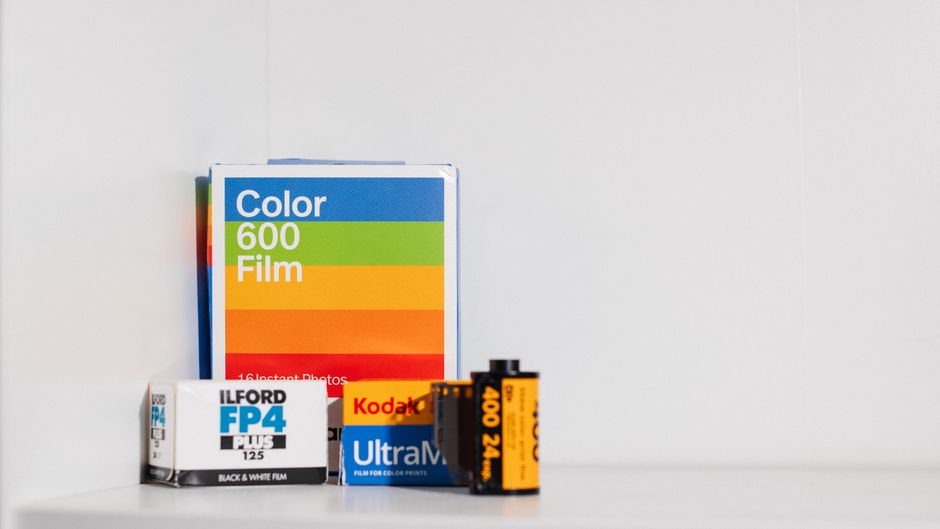 A collection of photographic and film development materials arranged on a light grey surface against a plain grey background. The items include a box of colour 600 film with a multicoloured label feat