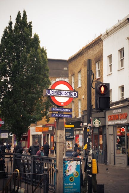 Nighttime view of the entrance to Bethnal Green Tube station, with illuminated signage and a large, grid-style glass window emitting warm yellow light from the station interior. The foreground feature